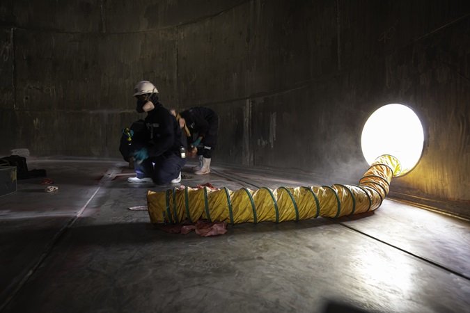 Fotografia scattata all'interno di una cisterna o silos con due lavoratori abbigliati per operare in spazi confinati e un sistema di aerazione forzata posizionato nell'ambiente.