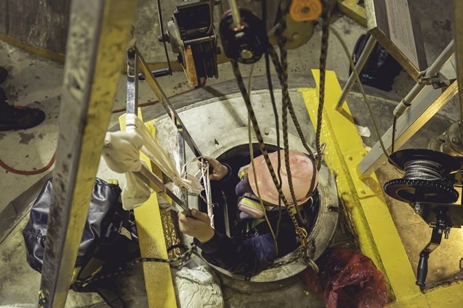 Vista dall'alto di un uomo fornito di tutti i DPI per operare in spazi confinati che esce da un passo d'uomo attraverso una scaletta a pioli, agganciato a un tripode per il recupero d'emergenza.