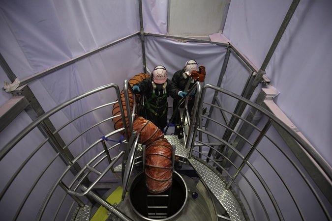 Fotografia dall'alto del punto di accesso a una cisterna o silos con inserito il sistema di aerazione forzata e due lavoratori in procinto di accedere allo spazio confinato.