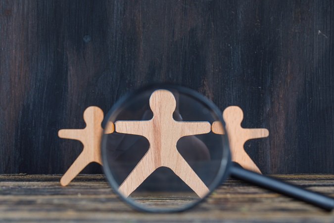 Tre omini di legno che rappresentano una squadra di lavoro. Uno di queste è sotto la lente d'ingrandimento per indicare il suo ruolo di preposto che richiede una formazione specifica: il corso preposto.