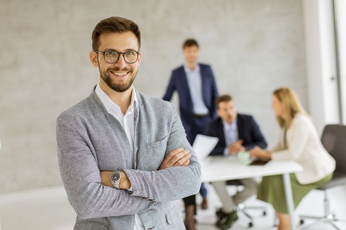 Un giovane uomo che sorride. Alle spalle tre persone intorno a un tavolo. L'uomo in primo piano rappresenta il capo, il datore di lavoro, da oggi soggetto a formazione obbligatoria per effetto del nuovo Accordo Stato Regioni.