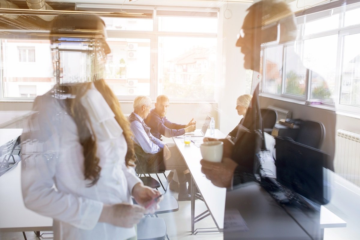 Riunione in un ufficio con 5 persone, riflessa attraverso una vetrata. La riunione periodica come momento in cui effettuare l'analisi dei dati e formalizzare la verifica dell'efficacia.