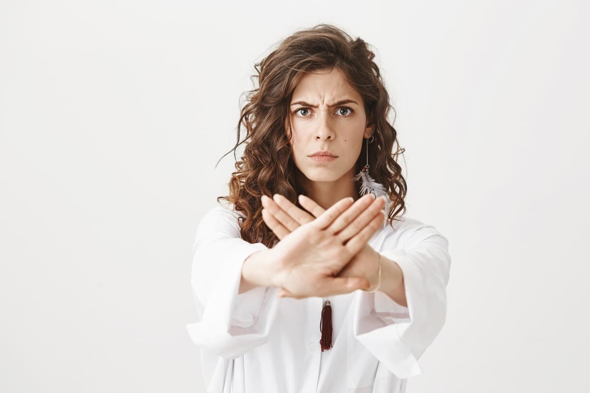 Una donna riccia vestita con una camicia bianca, stende le braccia di fronte a sé e forma una X con le mani. La sua espressione corrucciata e il gesto delle mani esprimono contrarietà, definiscono un limite: simbolo della indisponibilità alla corruzione.
